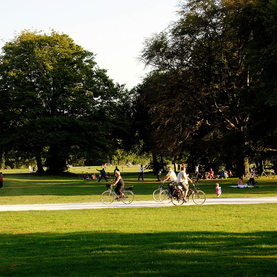 Lush park with green grass, tall trees, paved walkways, and people bustling about.