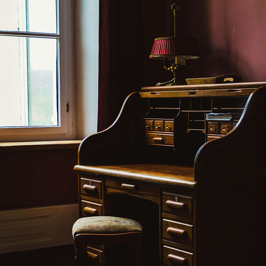 Elegant wood desk with lamp and stool next to large window.