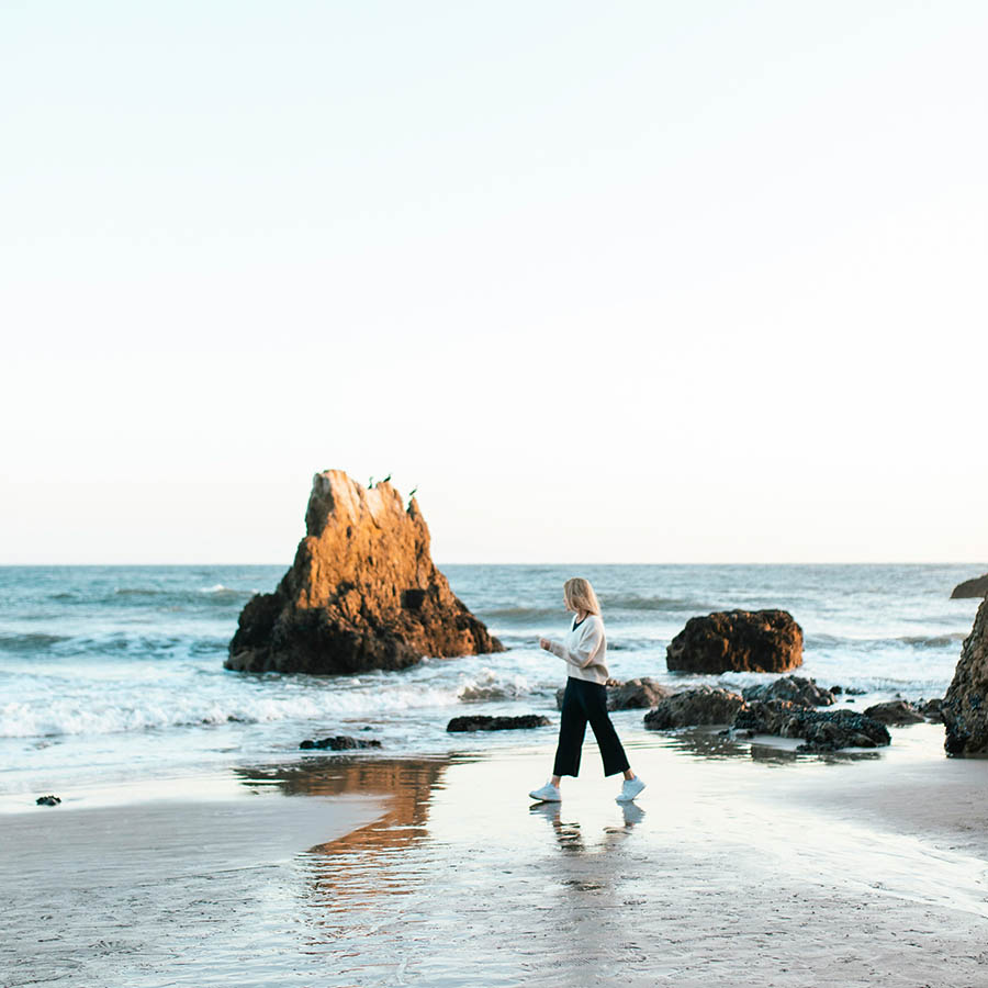 Person walking along sandy beach with towering rocks.