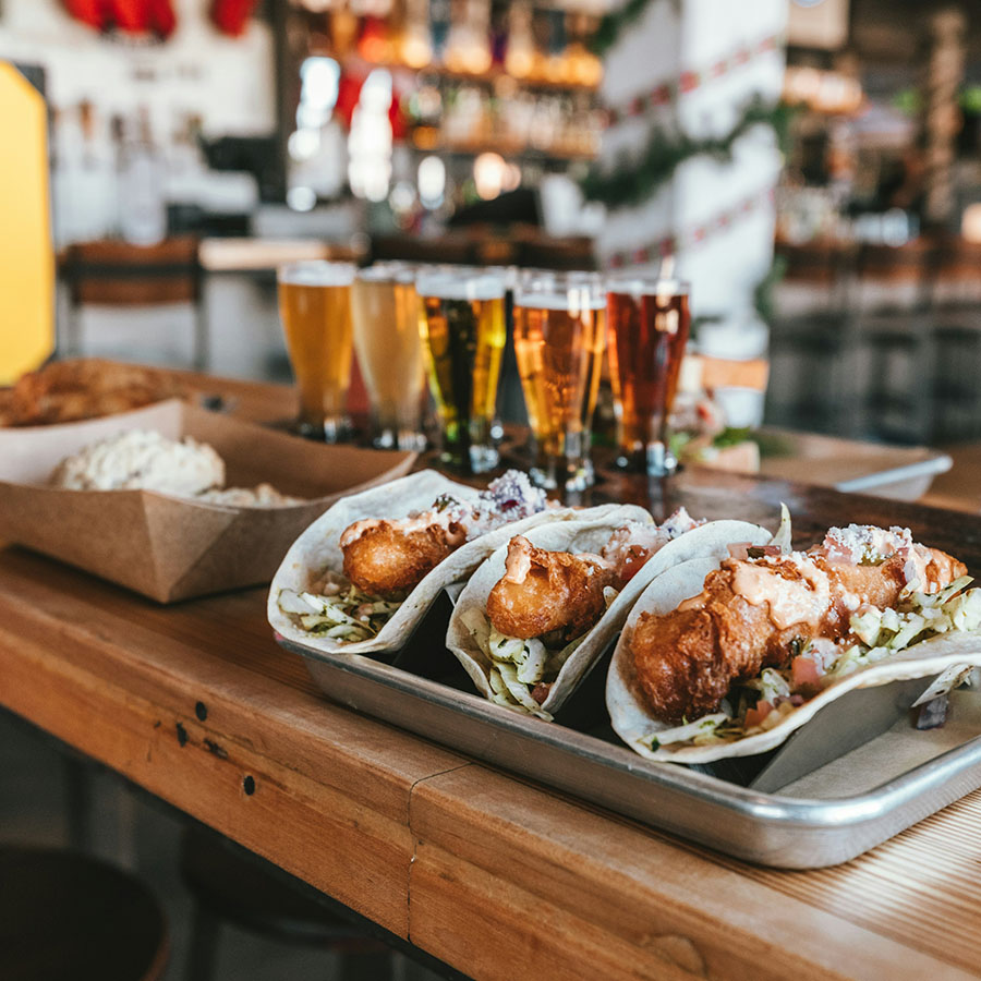 Flight of beers and plate of tacos on wood bar.