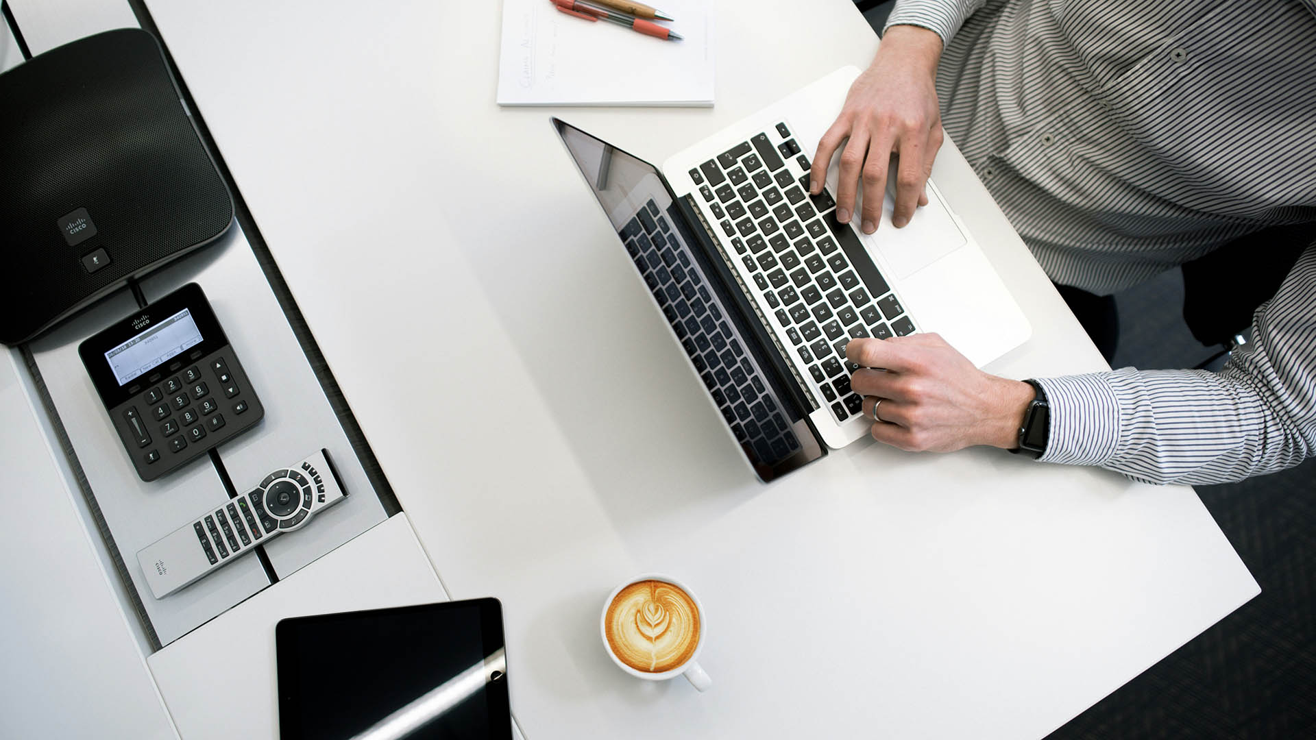 Person working at table with computer, tablet, notebook, and coffee mug.