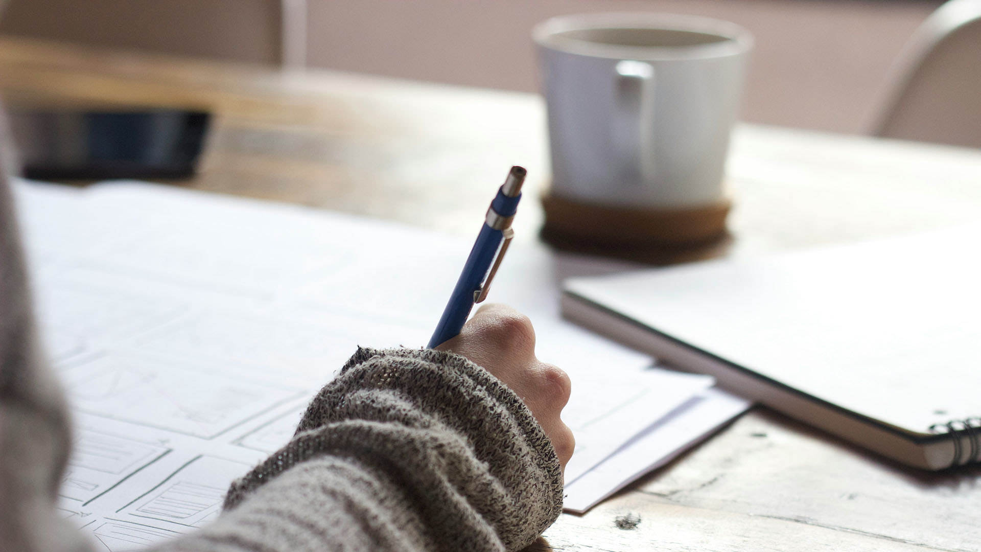 Person working at desk with papers, pencil, notebook, and coffee mug.